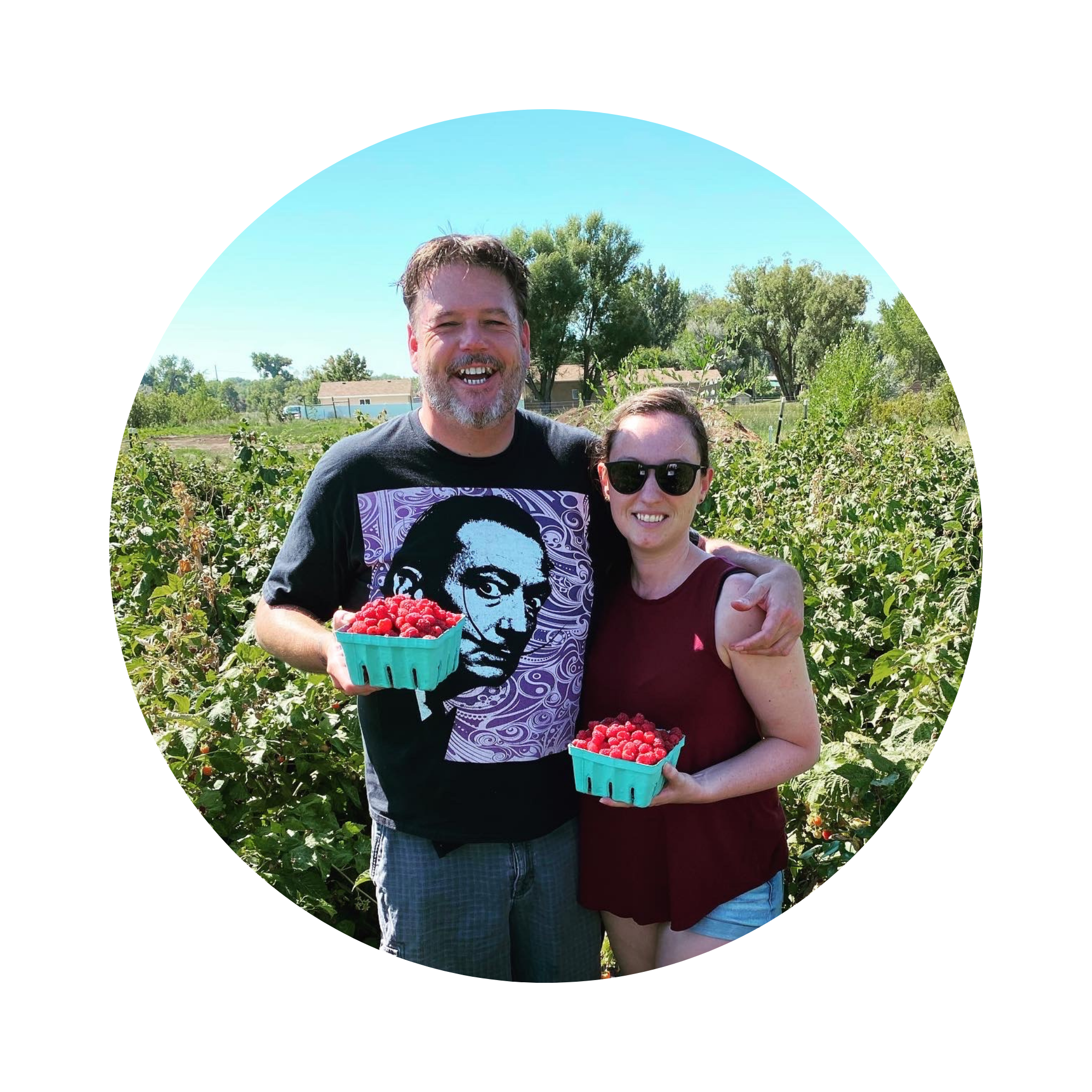 Crawford's Cottage owners, Phil and Kelli, holding containers of raspberries in a field. Phil is tall with short dark hair and a beard. Kelli comes up to his shoulder and is wearing a red shirt.